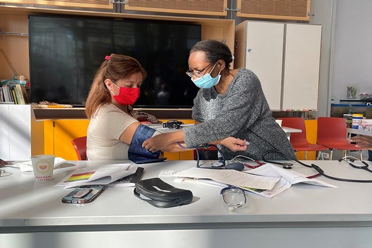 Woman stands while checking a seated woman's blood pressure 