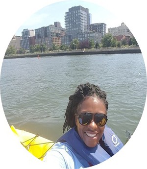 A woman dressed in blue vest and white leeves smiles into camera while a city and river sets in the backdrop