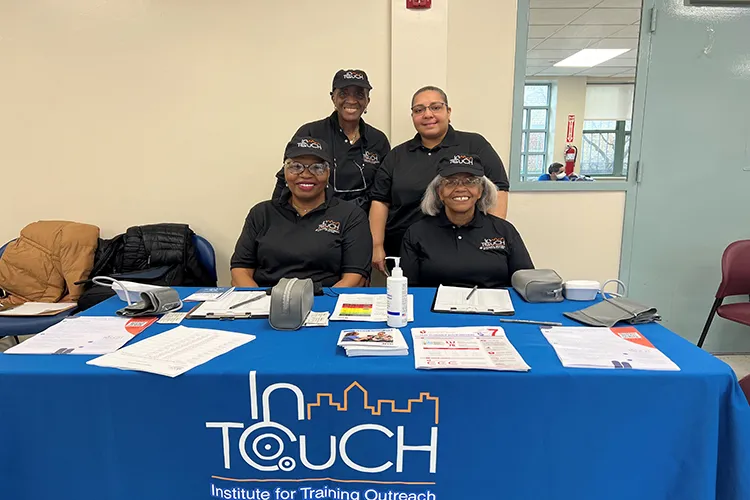 Four InTOuch members two seated and two standing smile into camera while seated behind a table covered with supplies and an InTOuch banner draped across the table