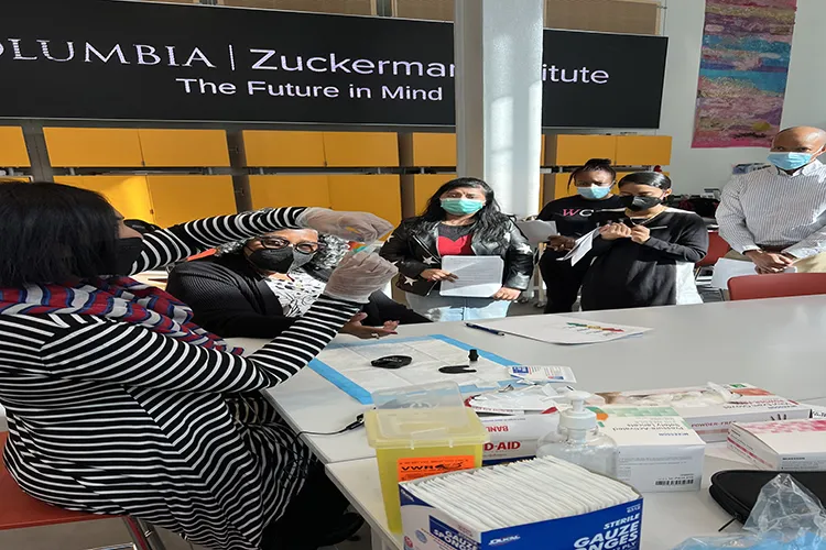 Dr. Mallaiah stands wearing a black and white striped top, plastic gloves and face maks to demonstrate blood glucose testing to a group of students standing around a table while wearing face masks.