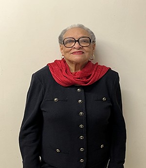 A woman dressed in dark rimmed eyeglasses wears a red shawl draped around her neck wears a dark blazer with gold and black buttons. She smiles into camera.