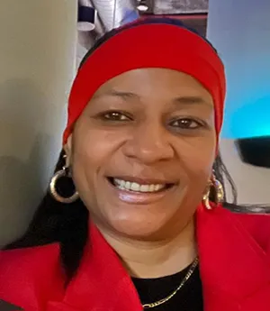 A woman smiles into wearing a red band across her head, gold earrings and chain. She has a black top on underneath a red blazer. 