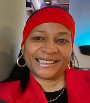 A woman smiles into wearing a red band across her head, gold earrings and chain. She has a black top on underneath a red blazer.