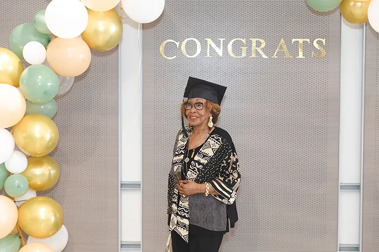 A woman dressed in a black and gold trimmed blouse and dark rimmed eyeglasses wears a black graduation cap while standing underneath a congrats signage and a colorful array of balloons adorn the wall