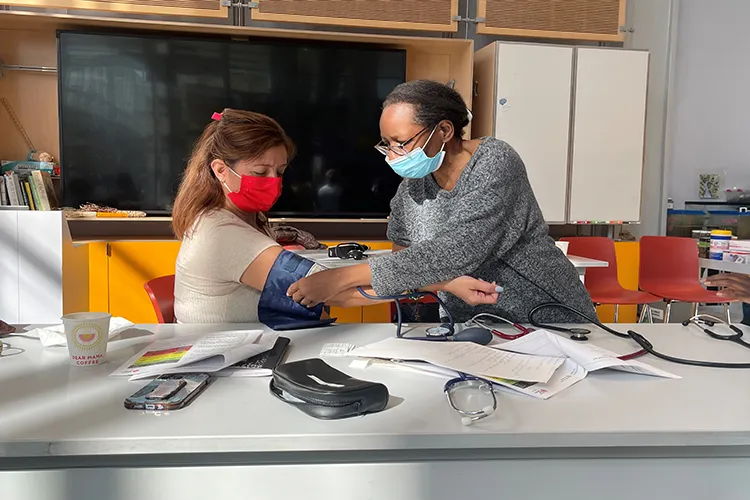Two women wear masks as one woman in eyeglasses stands to demonstrate how to check for blood pressure on the seated woman's arm.