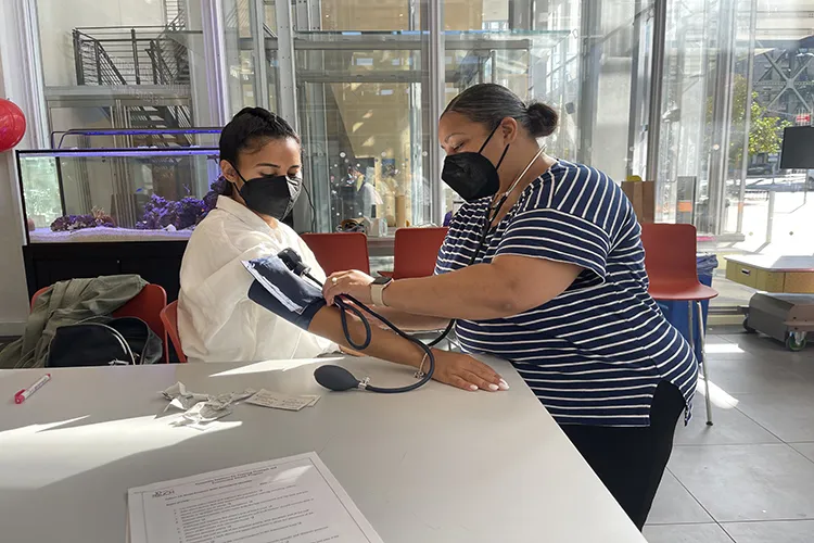 Two women wearing face masks demonstrate blood pressure checking as one woman stands to place the cuff around the arm of a seated womanin a white sweater
