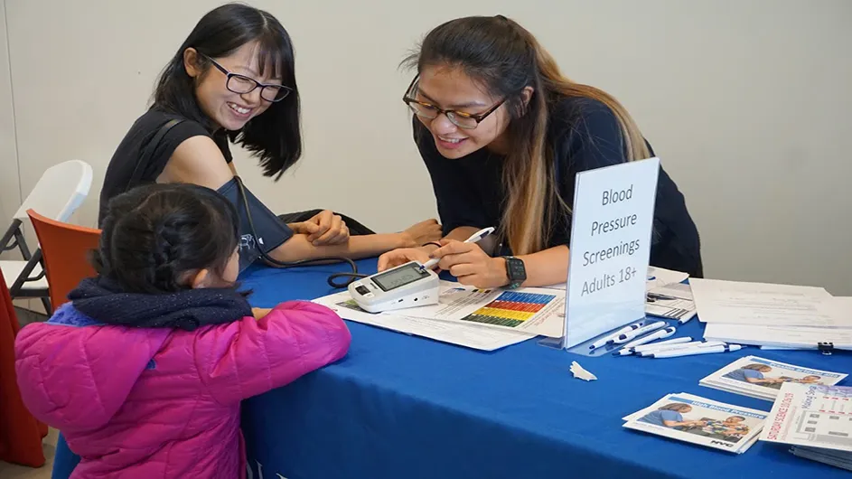 Wellness Center Staff Nurse, Cindy Hsu, takes a woman's blood pressure while explaining to the woman's daughter how the automatic blood pressure machine works.