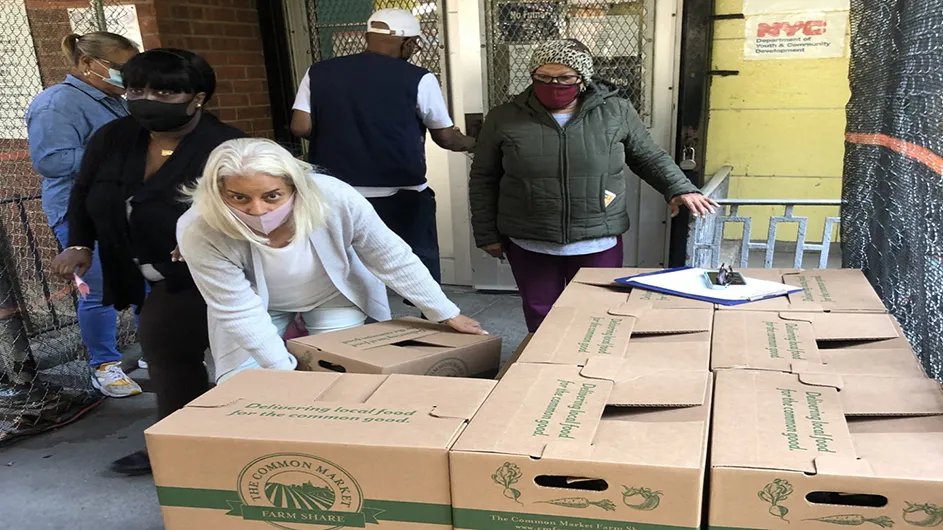 Stack of cardboard boxes with produce inside are being distributed by a staff member of the Jackie Robinson Senior Center. Seniors are seen in the background lined up to receive their farm share. 