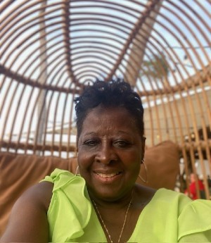Woman wears a neon lime green short sleeved blouse while seated in a bamboo chair. She wears a gold chain, earrings and smiles into camera.