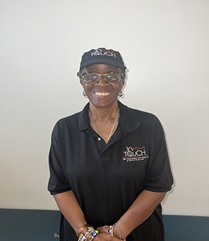 Woman stands in front of a beige wall while smiling into camera. She wears a dark InTOuch cap and matching polo shirt. She wears a tortoise shell framed eyeglasses with silver jewelry on her neck and earrings with muticolored bracelets.