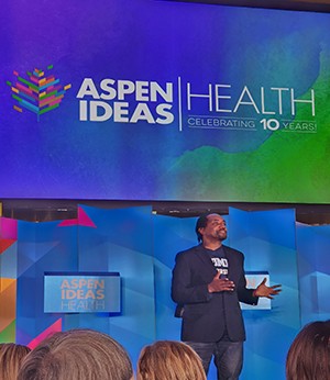 A man stands on stage in front of a projected slide that reads "Aspen Ideas, Health Celebrating 10 Years". He wears a dark blazer jacket and dark jeans while gestering with open hands to audience.