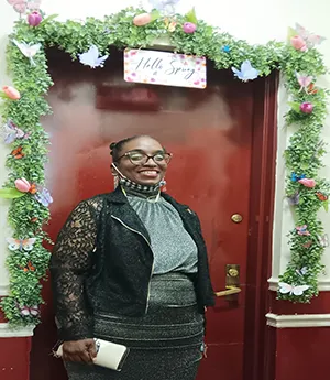 A woman stands in front of a red door wearing dark rimmed eyeglasses.  She smiles into camera wearing a grey dresses, dark jacket while smiling into camera. 