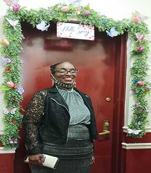 A woman stands in front of a red door wearing dark rimmed eyeglasses. She smiles into camera wearing a grey dresses, dark jacket while smiling into camera.